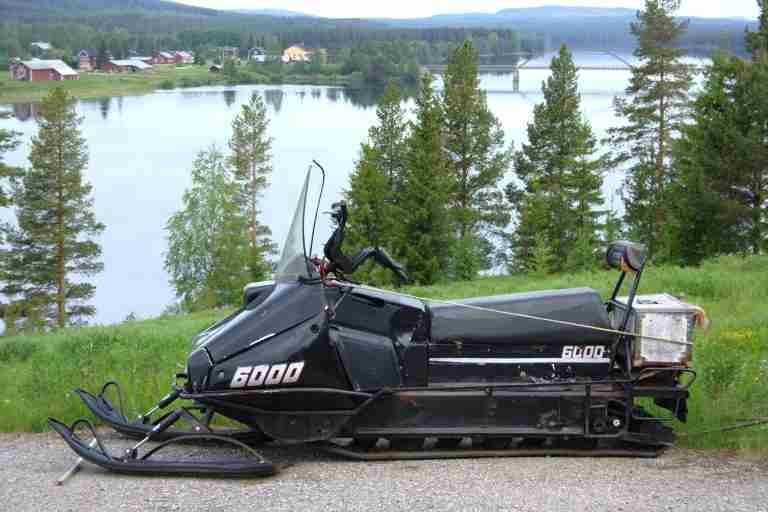 A snowmobile, financed with a 6000 loan despite bad credit, parked on the grass beside a tranquil lake with a backdrop of forest and scattered houses.
