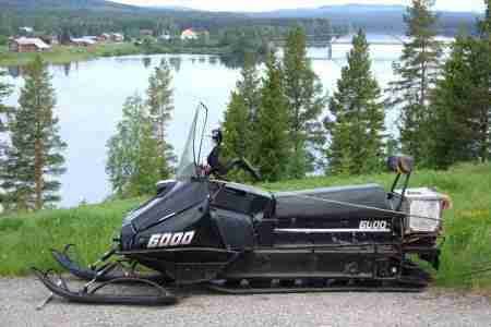 A snowmobile, financed with a 6000 loan despite bad credit, parked on the grass beside a tranquil lake with a backdrop of forest and scattered houses.