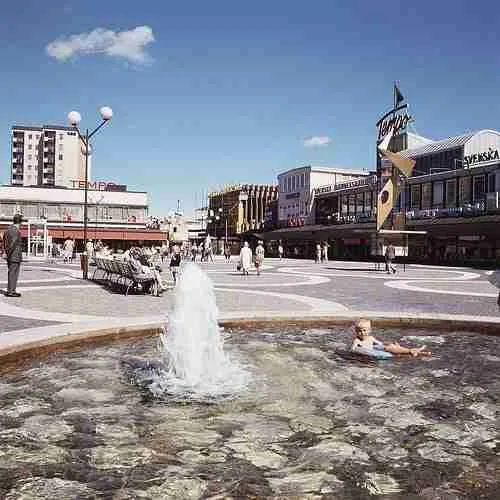 A sunny day at a city square with a fountain in the foreground and pedestrians walking by shops and cafes under a clear blue sky, discussing options for secured homeowner loans despite bad credit.