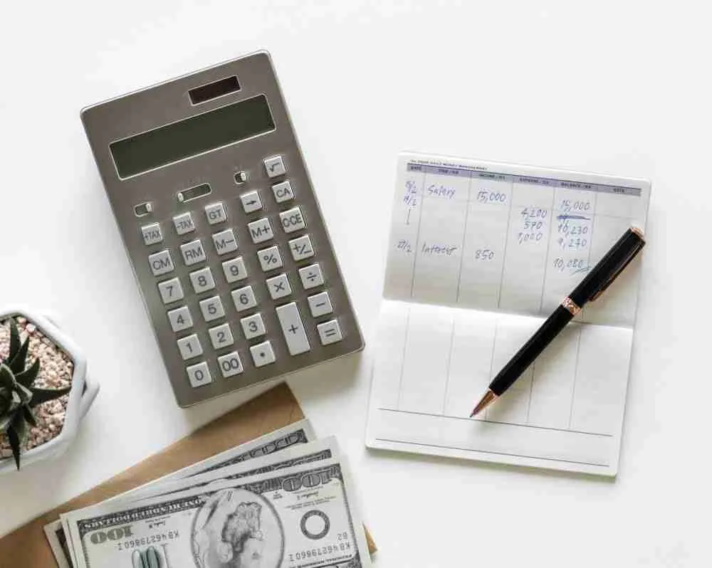 A desktop with financial planning tools: a calculator, a checkbook with written figures, a pen, and a batch of cash, suggesting budgeting or accounting activities.