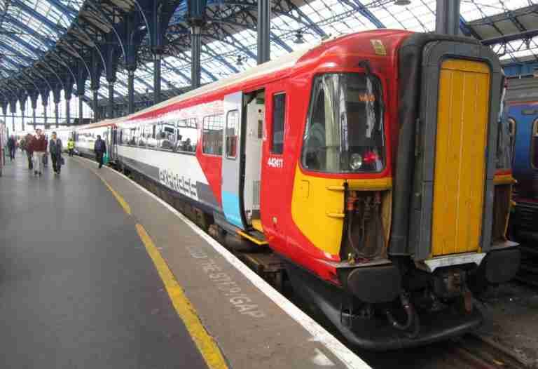 A red and white passenger train parked at a station platform with a blue steel and glass canopy, as travelers walk by.