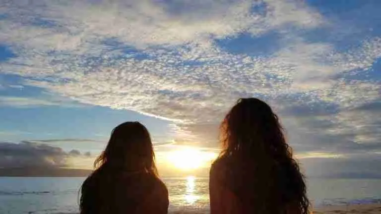 Two silhouetted figures enjoy a serene sunset over the ocean, as the sky above them is painted in a beautiful array of warm hues.
