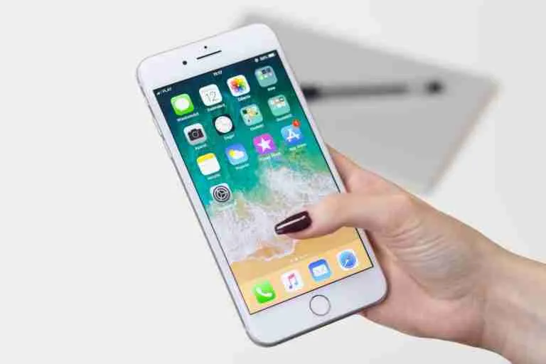A hand holding a smartphone with its home screen visible, displaying a variety of app icons against a wallpaper with a wave design.